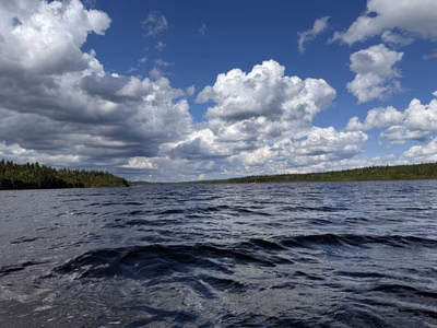 A lake bordered by forest beneath a blue sky filled with clouds.
